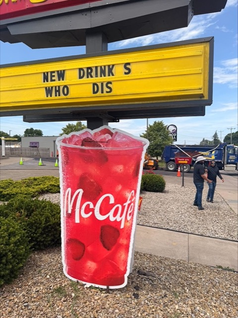 An oversized red McCafe beverage cup cutout staked beneath a NEW DRINKS WHO DIS marquee outside the restaurant