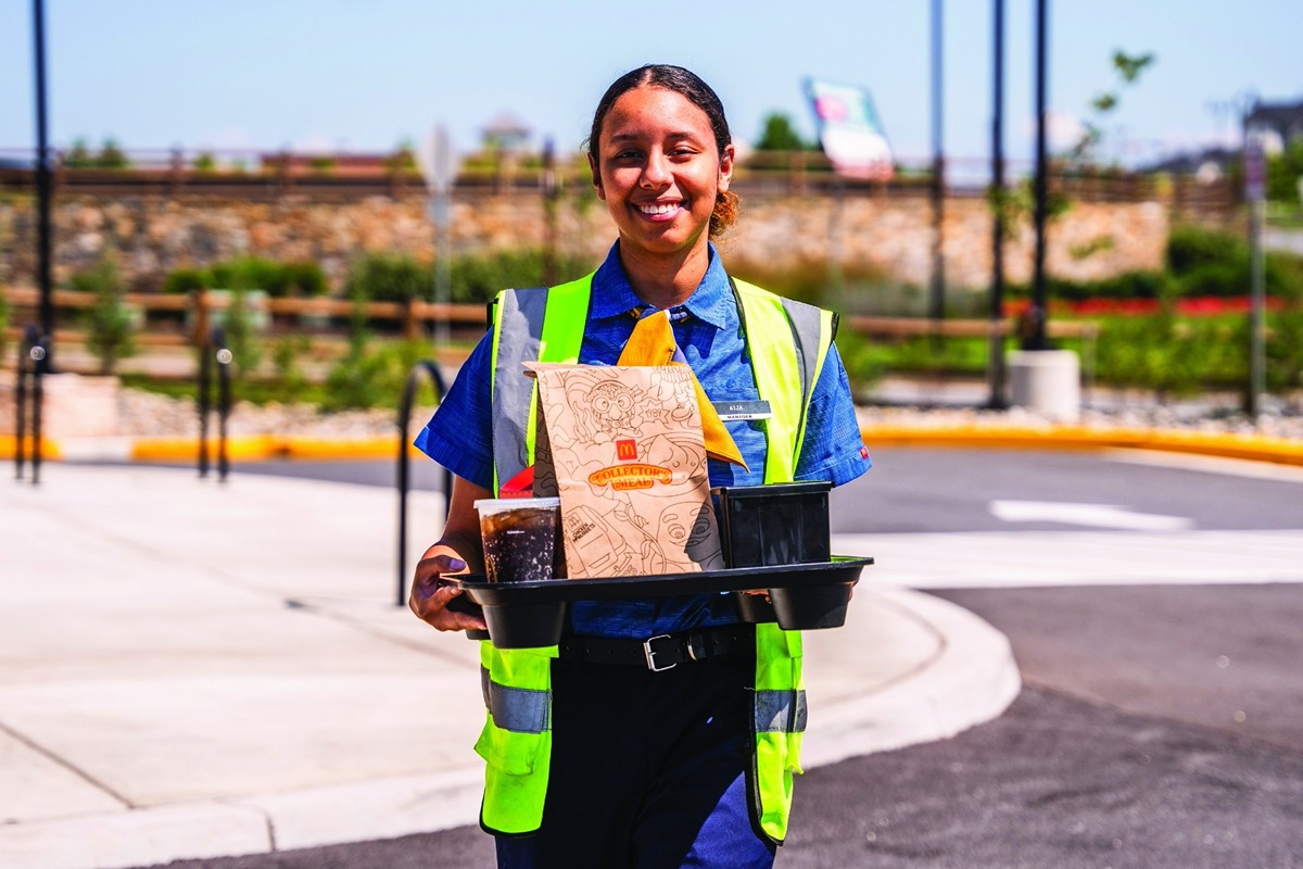 A McDonald's shift manager in a hi-vis vest carrying a curbside tray of drinks and a Collector's Meal bag out to a guest